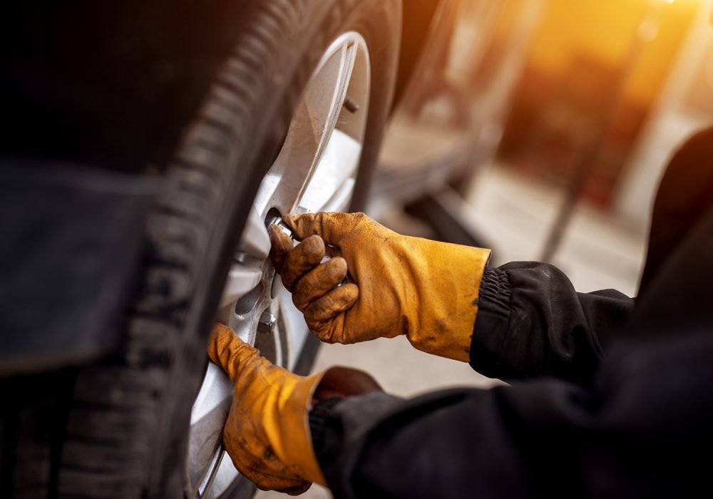 An experienced mechanic with orange gloves is putting screws on a placed wheel on a car.